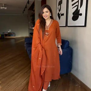 Peut inclure: Un ensemble tunique et pantalon de couleur rouille, à manches longues, avec des détails brodés. La tenue comprend une écharpe drapée et est portée avec des sandales transparentes. Le texte "Pink Wardrobe" est visible.