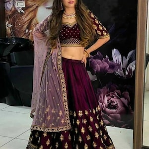 Puede incluir: Un atuendo formal de dos piezas en borgoña oscuro. La parte superior es una blusa corta de manga corta con bordados dorados. La falda es larga, con bordados a juego. Una bufanda transparente de color lavanda está drapeada sobre los hombros.