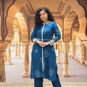 May include: A teal-colored, long-sleeved tunic and matching pants set. The tunic features white dot patterns and a white trim. The pants have a matching trim at the hem. The text "KRSURAT EVA" is visible on the tunic.