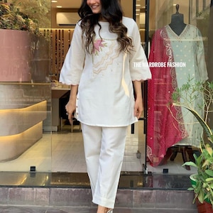 May include: A white two-piece outfit with a floral embroidered design on the top. The outfit includes a tunic with three-quarter sleeves and matching pants. The text "THE WARDROBE FASHION" is visible on the glass.