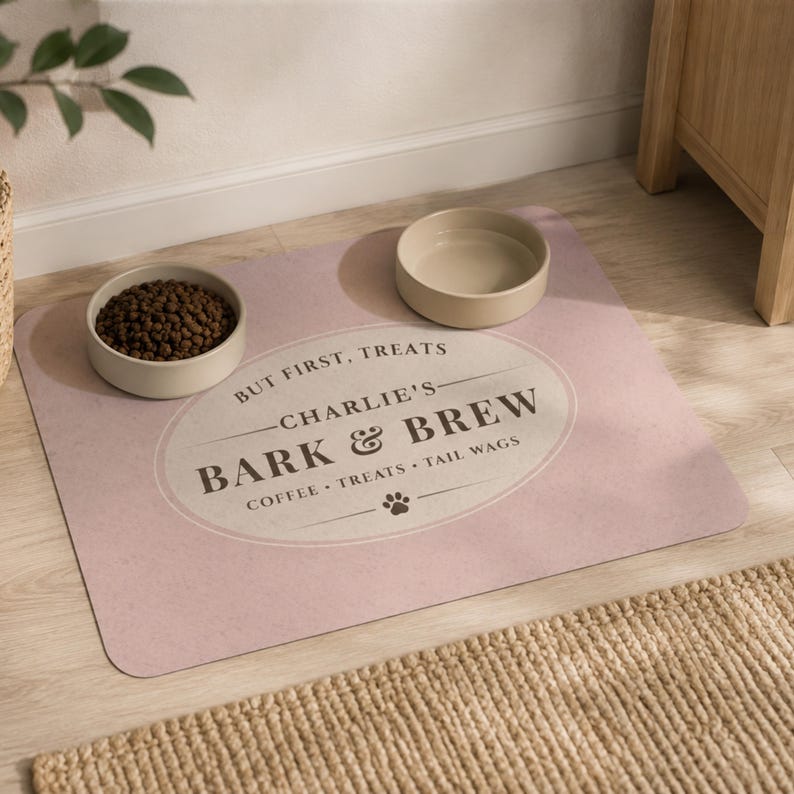 May include: A rectangular light pink pet feeding mat featuring a central oval design with the text "BUT FIRST, TREATS", "CHARLIE'S BARK & BREW", and "COFFEE &bull; TREATS &bull; TAIL WAGS" with a paw print icon. Two beige ceramic bowls sit on top of the mat.