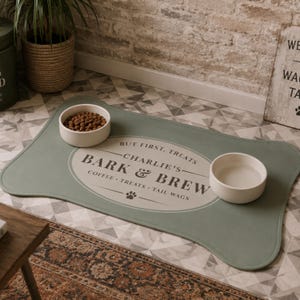 May include: A sage green pet feeding mat with a bone-shaped outline and a central oval graphic. The text reads: BUT FIRST, TREATS, CHARLIE'S, BARK & BREW, COFFEE, TREATS, TAIL WAGS. Two white ceramic bowls sit on the mat, one filled with brown kibble.