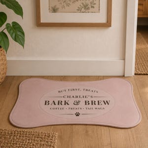 May include: A bone-shaped mat in a soft dusty pink color featuring a dark brown oval graphic with the text "BUT FIRST, TREATS", "CHARLIE'S", "BARK & BREW", and "COFFEE &bull; TREATS &bull; TAIL WAGS" along with a small paw print icon centered at the bottom.