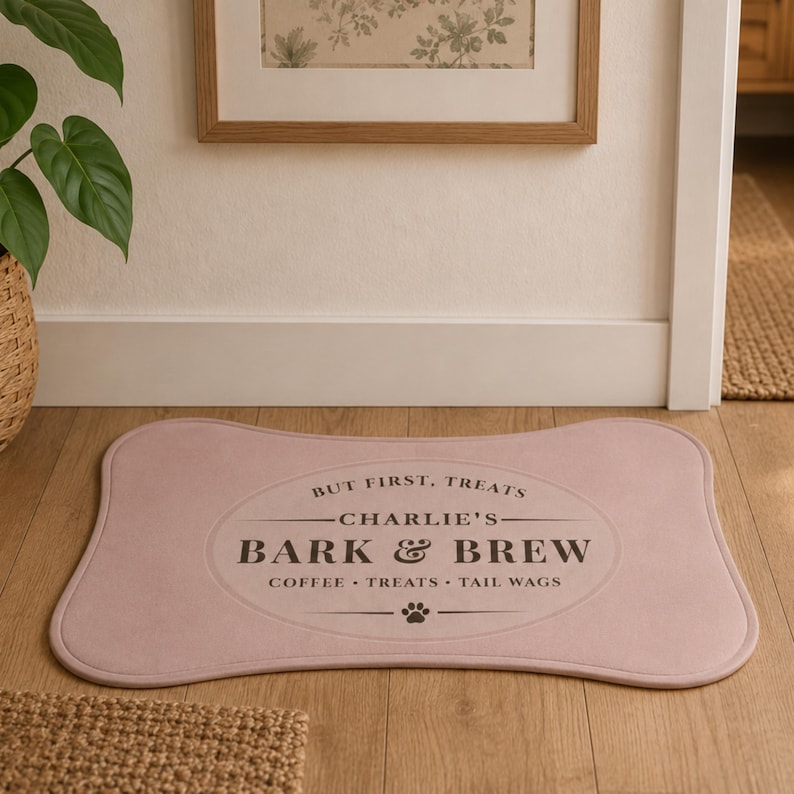 May include: A bone-shaped mat in a soft dusty pink color featuring a dark brown oval graphic with the text "BUT FIRST, TREATS", "CHARLIE'S", "BARK & BREW", and "COFFEE &bull; TREATS &bull; TAIL WAGS" along with a small paw print icon centered at the bottom.
