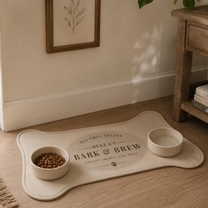 May include: A bone-shaped beige pet feeding mat with two round off-white bowls. The mat features a central oval design with the text "BUT FIRST, TREATS", "BELLA'S", "BARK & BREW", "COFFEE &bull; TREATS &bull; TAIL WAGS" and a small paw print icon.