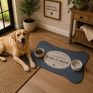 May include: A bone-shaped dark blue pet feeding mat featuring a central oval design with the text "BUT FIRST, TREATS", "CHARLIE'S", "BARK & BREW", and "COFFEE &bull; TREATS &bull; TAIL WAGS" with a paw print icon. Two white ceramic bowls sit on the mat.