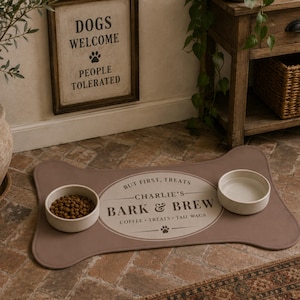 May include: A bone-shaped brown pet feeding mat featuring a central oval design with the text "BUT FIRST, TREATS", "CHARLIE'S BARK & BREW", and "COFFEE &bull; TREATS &bull; TAIL WAGS" with two round off-white bowls, one filled with dry kibble and one with water.