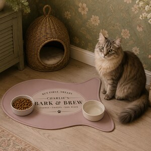 May include: A dusty rose, fish-shaped silicone pet feeding mat featuring the printed text "BUT FIRST, TREATS", "CHARLIE'S BARK & BREW", and "COFFEE &bull; TREATS &bull; TAIL WAGS" with a small paw print icon, holding two white ceramic bowls for food and water.