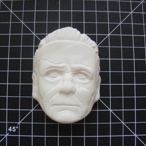May include: A white plaster mold of a person's face. The face has wrinkles and lines, and the eyes are closed. The mold is on a black and white grid background.