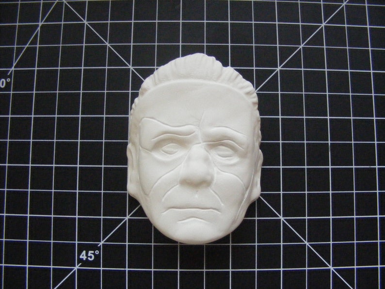May include: A white plaster mold of a person's face. The face has wrinkles and lines, and the eyes are closed. The mold is on a black and white grid background.