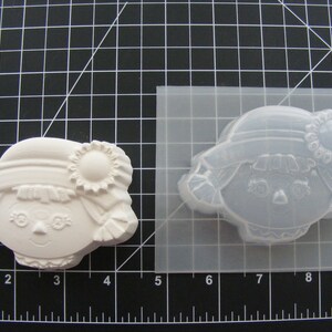 May include: A white plastic mold for making a small doll with a hat and a flower. The mold is on a black grid background.