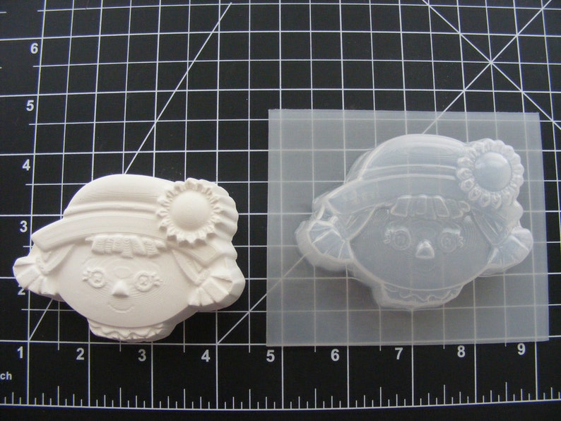 May include: A white plastic mold for making a small doll with a hat and a flower. The mold is on a black grid background.
