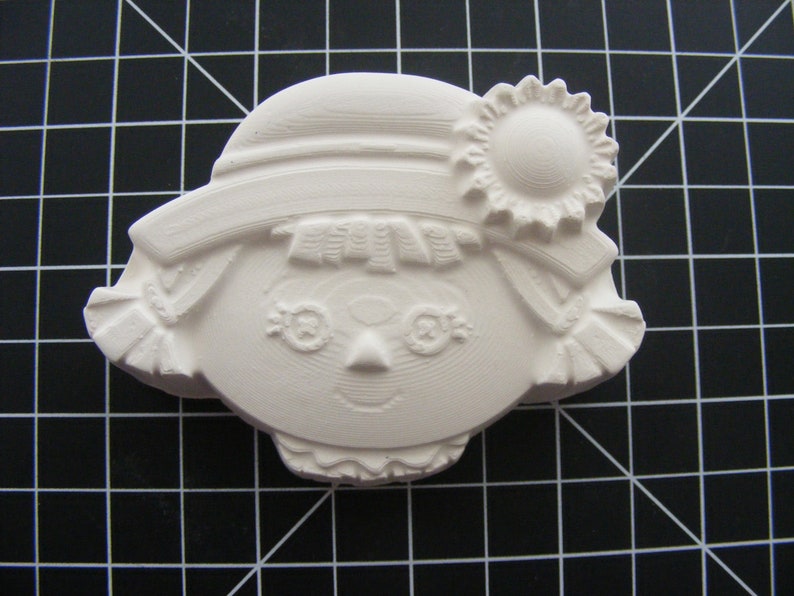 May include: White ceramic figurine of a girl with a hat and a flower. The girl has a simple face with eyes, a nose, and a smile. The figurine is a blank canvas for painting.