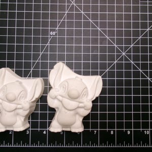 May include: Two unpainted Stitch figurines, a popular cartoon character, are displayed on a black cutting mat with a grid pattern. The figurines are white and feature Stitch's distinctive large ears and facial features. The mat has inch measurements.