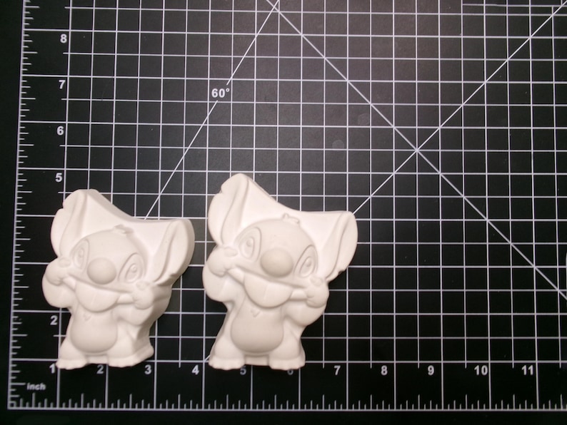 May include: Two unpainted Stitch figurines, a popular cartoon character, are displayed on a black cutting mat with a grid pattern. The figurines are white and feature Stitch's distinctive large ears and facial features. The mat has inch measurements.