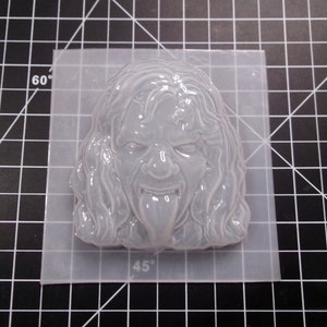 May include: A white silicone mold of a face with long hair and a tongue sticking out. The mold is on a clear plastic mat with markings for 45 and 60 degrees.