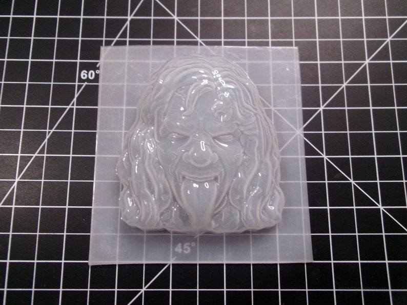 May include: A white silicone mold of a face with long hair and a tongue sticking out. The mold is on a clear plastic mat with markings for 45 and 60 degrees.