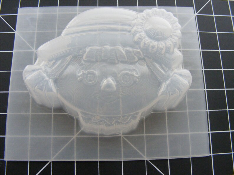 May include: Clear plastic mold for making a doll-shaped soap or candle. The mold features a girl with a hat and a flower on her head.