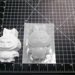 Puede incluir: Una figura blanca con forma de conejo y un molde de silicona transparente. La figura tiene una cara detallada y una textura similar a la piel. El molde tiene la forma de la figura. Los objetos est&aacute;n sobre una esterilla de corte negra con medidas en cm.