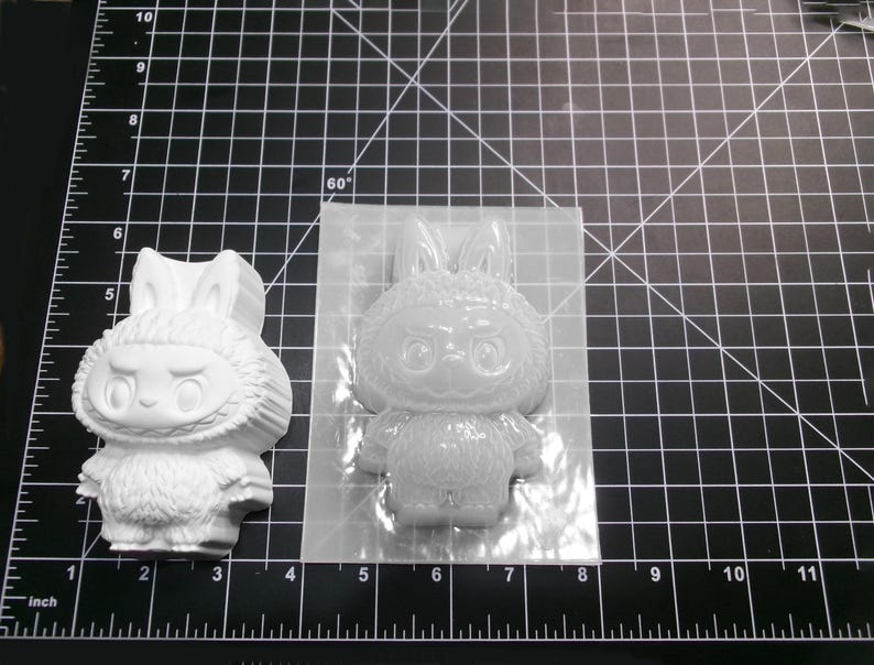 Puede incluir: Una figura blanca con forma de conejo y un molde de silicona transparente. La figura tiene una cara detallada y una textura similar a la piel. El molde tiene la forma de la figura. Los objetos est&aacute;n sobre una esterilla de corte negra con medidas en cm.