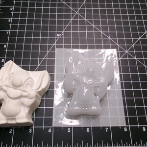 May include: A white Stitch figurine and a clear silicone mold of Stitch are displayed on a black cutting mat. The figurine is a solid, three-dimensional form, while the mold is transparent and flexible, showing the character's features.