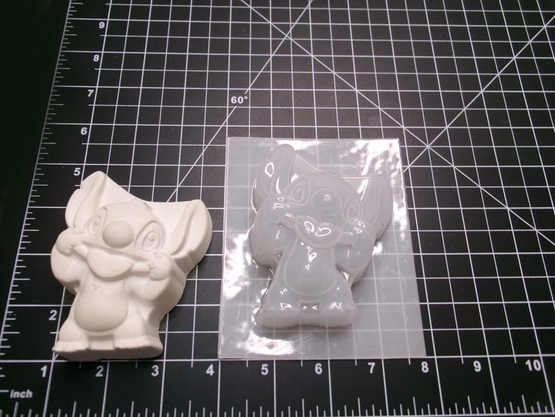 May include: A white Stitch figurine and a clear silicone mold of Stitch are displayed on a black cutting mat. The figurine is a solid, three-dimensional form, while the mold is transparent and flexible, showing the character's features.