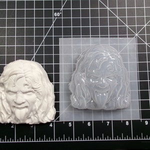 May include: A white silicone mold of a person's face with long hair and a tongue sticking out. The mold is on a black and white grid background.