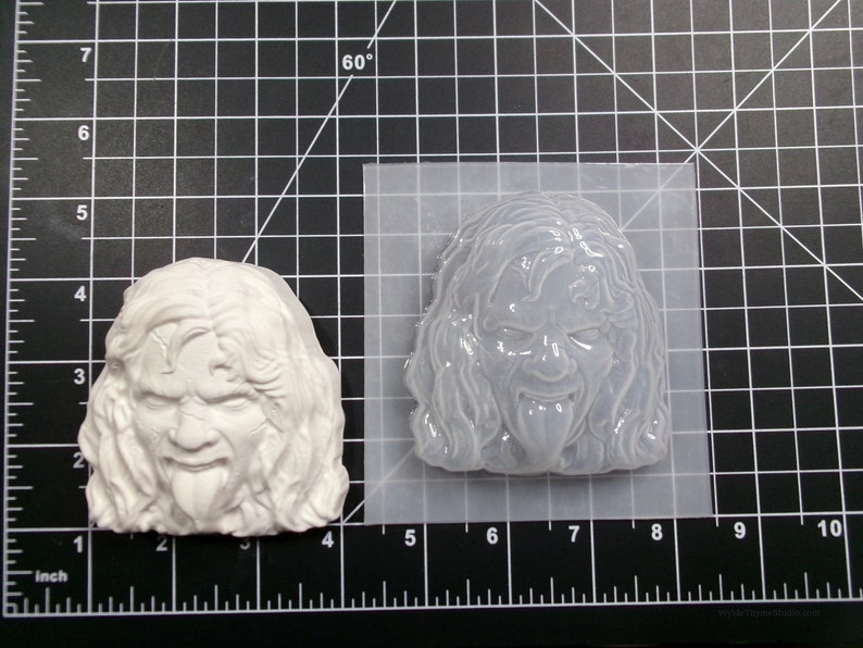 May include: A white silicone mold of a person's face with long hair and a tongue sticking out. The mold is on a black and white grid background.