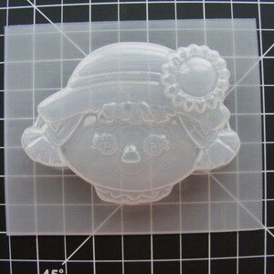 May include: A clear plastic mold for making a resin doll head. The mold features a detailed face with eyes, nose, and mouth, as well as a hat and flower.