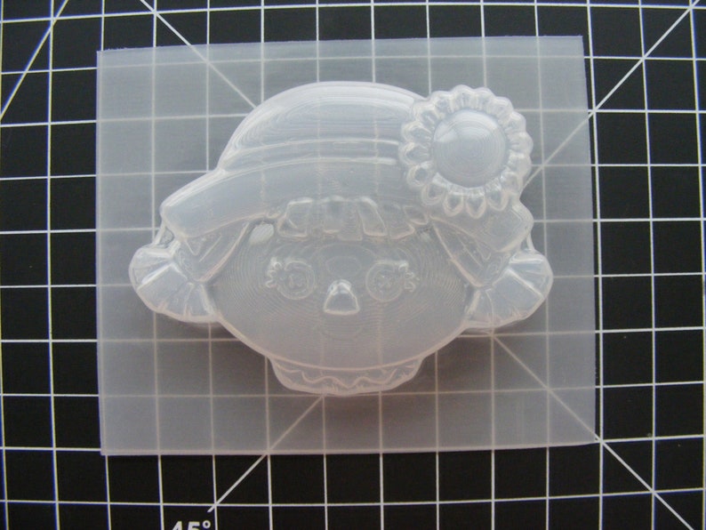 May include: A clear plastic mold for making a resin doll head. The mold features a detailed face with eyes, nose, and mouth, as well as a hat and flower.