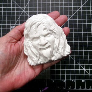 May include: White plaster sculpture of a person's face with long hair and a tongue sticking out.