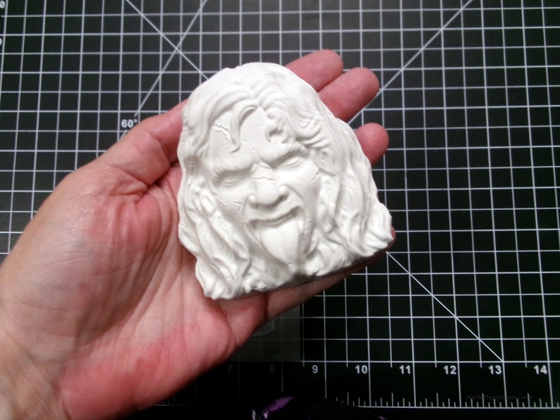 May include: White plaster sculpture of a person's face with long hair and a tongue sticking out.