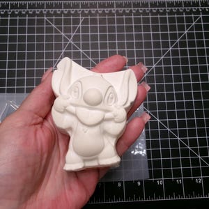 May include: A light-colored, unpainted Stitch figurine, held in a hand. The figurine is a cartoon character with large ears, a round nose, and a wide grin. The figurine is made of a smooth material. The background is a black cutting mat.