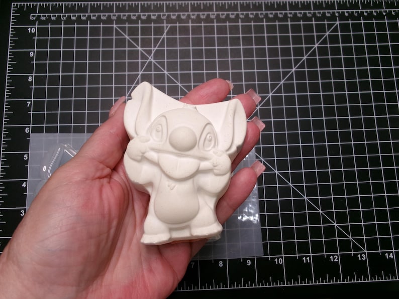 May include: A light-colored, unpainted Stitch figurine, held in a hand. The figurine is a cartoon character with large ears, a round nose, and a wide grin. The figurine is made of a smooth material. The background is a black cutting mat.