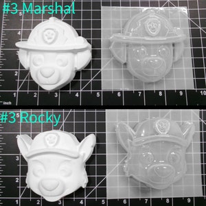May include: Two white plastic molds for making Paw Patrol character figurines. The molds are on a gridded surface. The molds are labeled #3 Marshal and #3 Rocky.