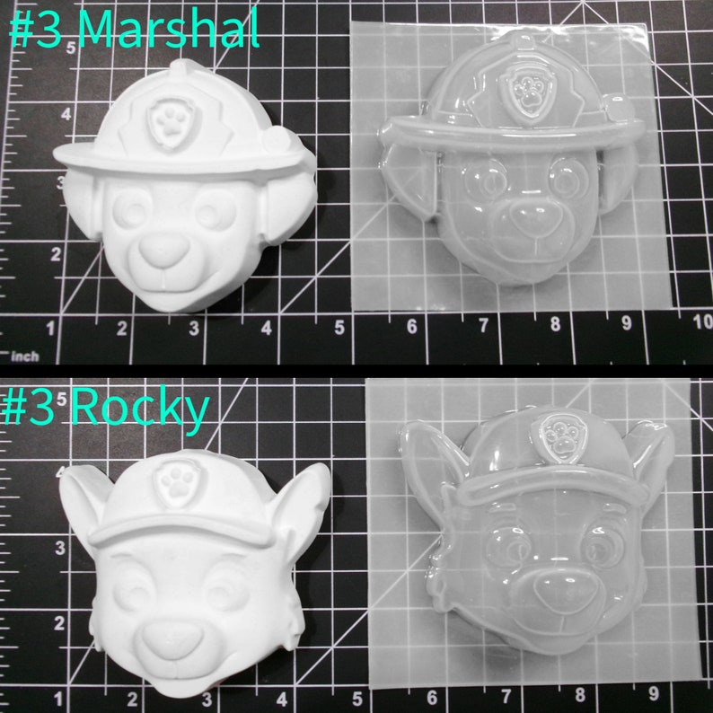 May include: Two white plastic molds for making Paw Patrol character figurines. The molds are on a gridded surface. The molds are labeled #3 Marshal and #3 Rocky.