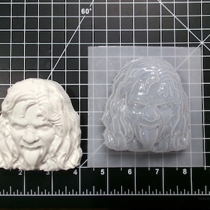 May include: A white silicone mold of a person's face with long hair and a tongue sticking out. The mold is on a black grid background.