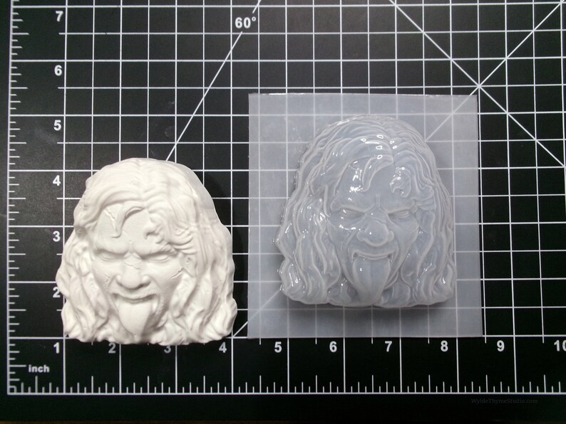 May include: A white silicone mold of a person's face with long hair and a tongue sticking out. The mold is on a black grid background.