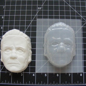 May include: A white plastic mold of a man's face. The mold is approximately 4 inches wide and 3 inches tall. The mold is used to create a replica of the face.