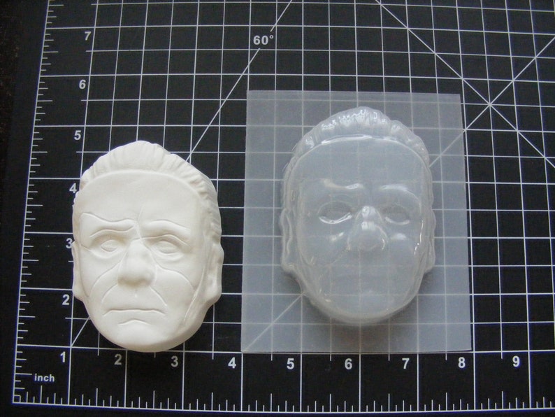 May include: A white plastic mold of a man's face. The mold is approximately 4 inches wide and 3 inches tall. The mold is used to create a replica of the face.