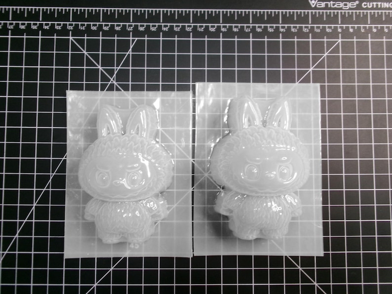 Puede incluir: Dos moldes transparentes con forma de conejito se muestran sobre una esterilla de corte negra. Cada molde est&aacute; encerrado en una l&aacute;mina de pl&aacute;stico transparente. El dise&ntilde;o del conejito presenta orejas grandes, una cara redonda y un cuerpo peque&ntilde;o.