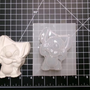 May include: A white, three-dimensional Stitch figure stands next to a clear silicone mold of the same character. The mold is on a black grid surface with inch measurements.