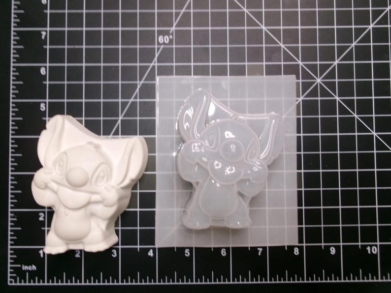 May include: A white, three-dimensional Stitch figure stands next to a clear silicone mold of the same character. The mold is on a black grid surface with inch measurements.