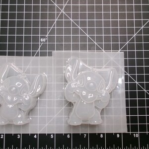 May include: Two clear silicone molds featuring a cartoon character with large ears and a friendly expression. The molds are on a grid-marked surface, with measurements in inches visible along the bottom edge. The molds are for crafting.