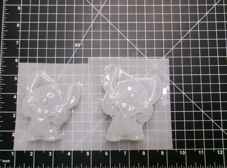 May include: Two clear silicone molds featuring a cartoon character with large ears and a friendly expression. The molds are on a grid-marked surface, with measurements in inches visible along the bottom edge. The molds are for crafting.