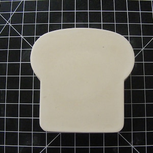 Op de afbeelding: Een witte keramische broodvorm in de vorm van een brood. De vorm staat op een zwart rooster.