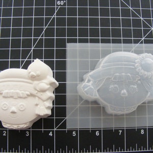 May include: A white plastic mold of a girl with a hat and a flower. The mold is on a black grid background.
