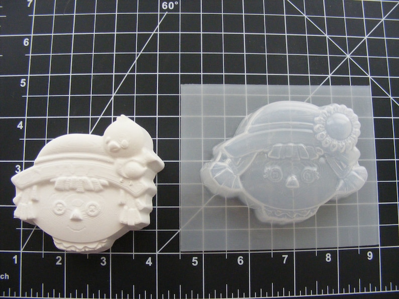 May include: A white plastic mold of a girl with a hat and a flower. The mold is on a black grid background.
