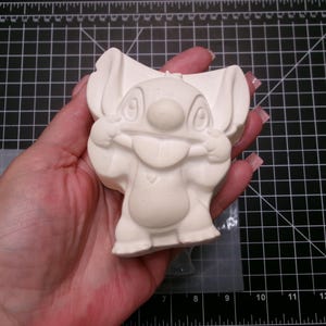May include: A light beige Stitch figurine, held in a hand, is displayed against a black grid background. The figurine has large ears, a wide smile, and is in a playful pose. The figurine is made of a smooth, matte material.