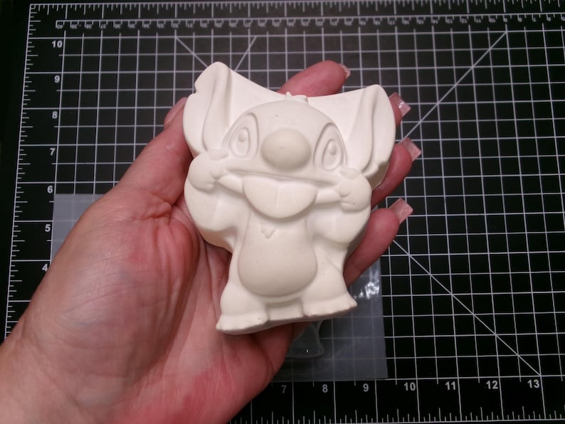 May include: A light beige Stitch figurine, held in a hand, is displayed against a black grid background. The figurine has large ears, a wide smile, and is in a playful pose. The figurine is made of a smooth, matte material.